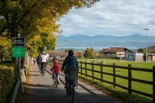 Family cycling together on a leafy community bike path in Épagny Metz-Tessy, with sports fields, hills, and the distant blue of Lake Annecy under soft autumn light.