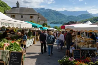 Open-air village market in Haute-Savoie with vendors, fresh produce, local shoppers, and mountains in the background under soft morning light