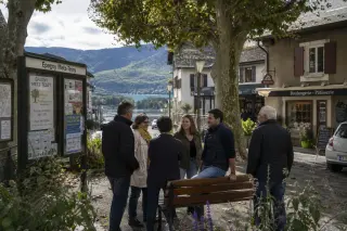 Residents gather around a bench in Épagny Metz-Tessy village square, discussing near civic bulletin boards, with trees, shopfronts, and glimpses of hills and lake in autumn light.