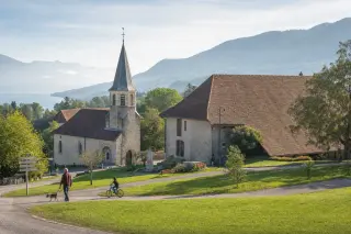 Historic village square with old stone buildings, tiled roofs, gentle morning light, wooded hills and lake in the background, locals casually passing by.