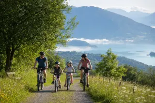 Family with two children cycling on a green path near Épagny Metz-Tessy, wildflowers along the trail, Lake Annecy and wooded hills in the background, morning light and peaceful atmosphere.