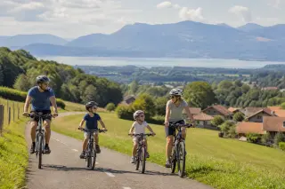 Family cycling along a bike path near Épagny Metz-Tessy with meadows, wooded patches, distant hills, and tiled-roof farmhouses in late summer light