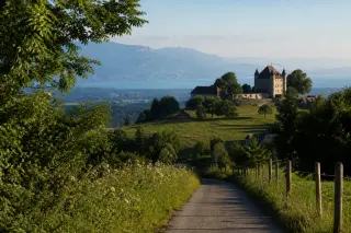Château de Montrottier overlooks a green meadow from a wooded hillside under soft, late afternoon light, with distant views of the Annecy basin.
