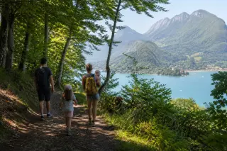Family walking on a leafy trail through woods overlooking Lake Annecy in the morning light, with glimpses of the lake and mountains in the background.