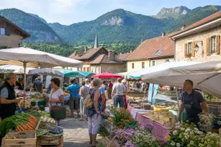 Outdoor village market in Haute-Savoie with people, local stalls, mountain hills in the background, and soft natural summer light.