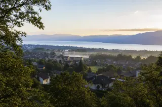 Panoramic view of Épagny Metz-Tessy rooftops with Lake Annecy and forested hills in soft morning light, subtle mist above the landscape.