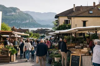 Morning outdoor market scene in a Haute-Savoie village with locals among wooden stalls, village homes, and gentle alpine hills in the background