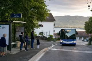 Residents waiting for a bus at a village stop in Épagny Metz-Tessy with tiled rooftops, soft green hills, and a glimpse of lake and mountains in the background on a calm, early morning.